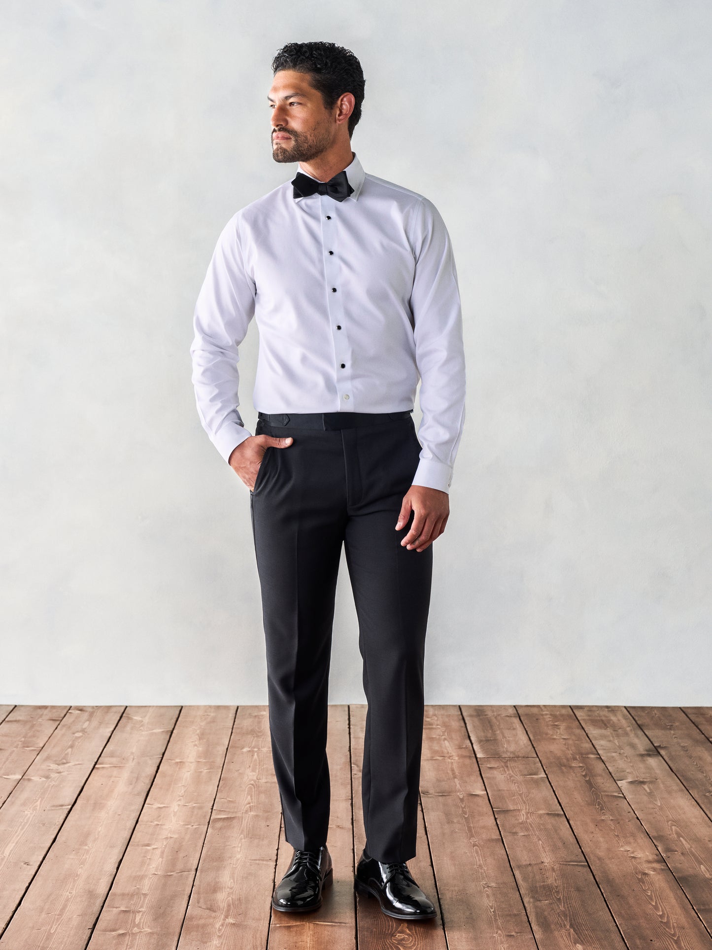 Man wearing black dress pants and a white dress shirt, hands in pockets, standing on wooden floorboards with a light gray background.