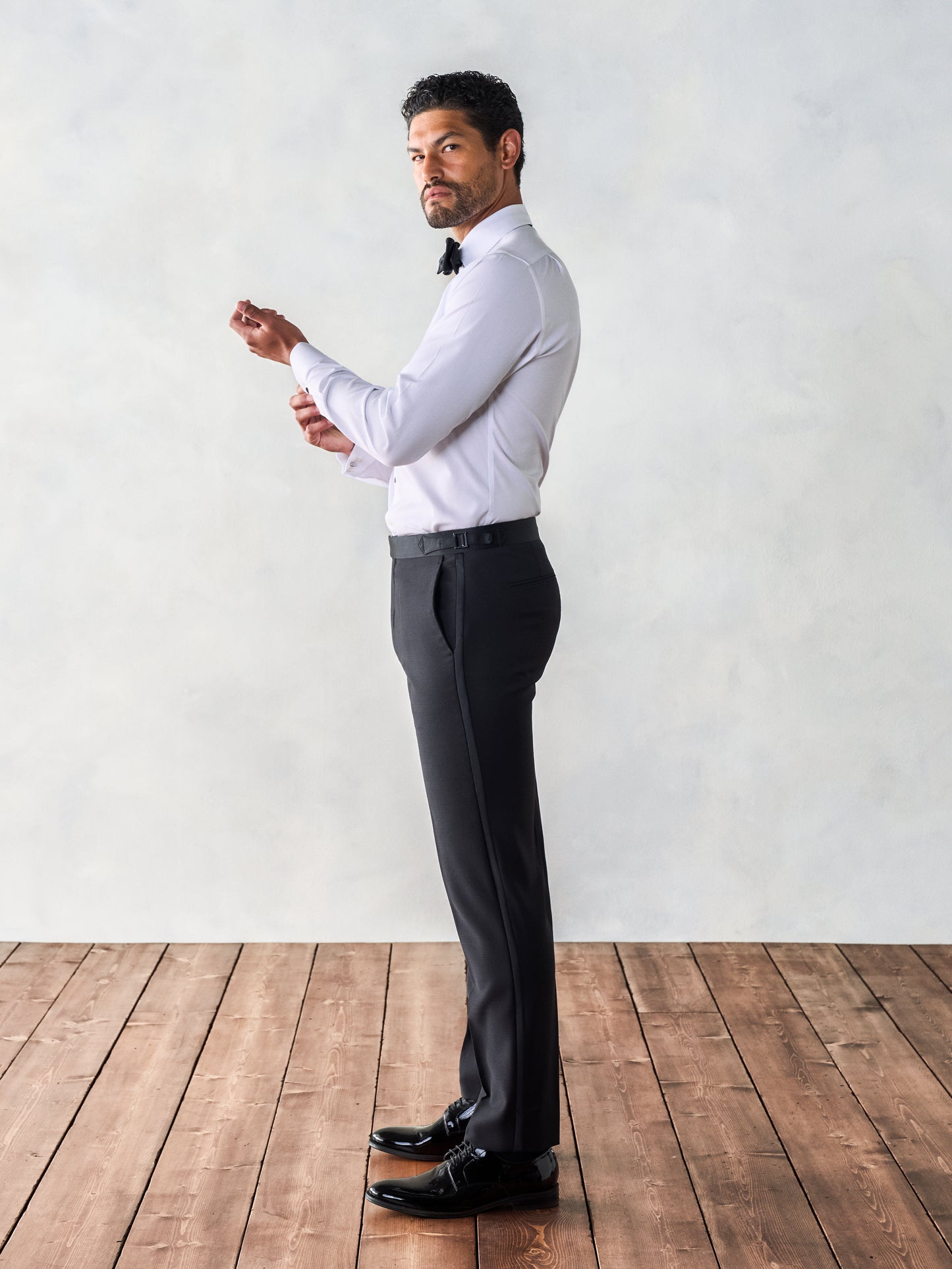 Side view of a man adjusting his cufflinks while wearing black dress pants and a white shirt, standing on wooden floorboards.