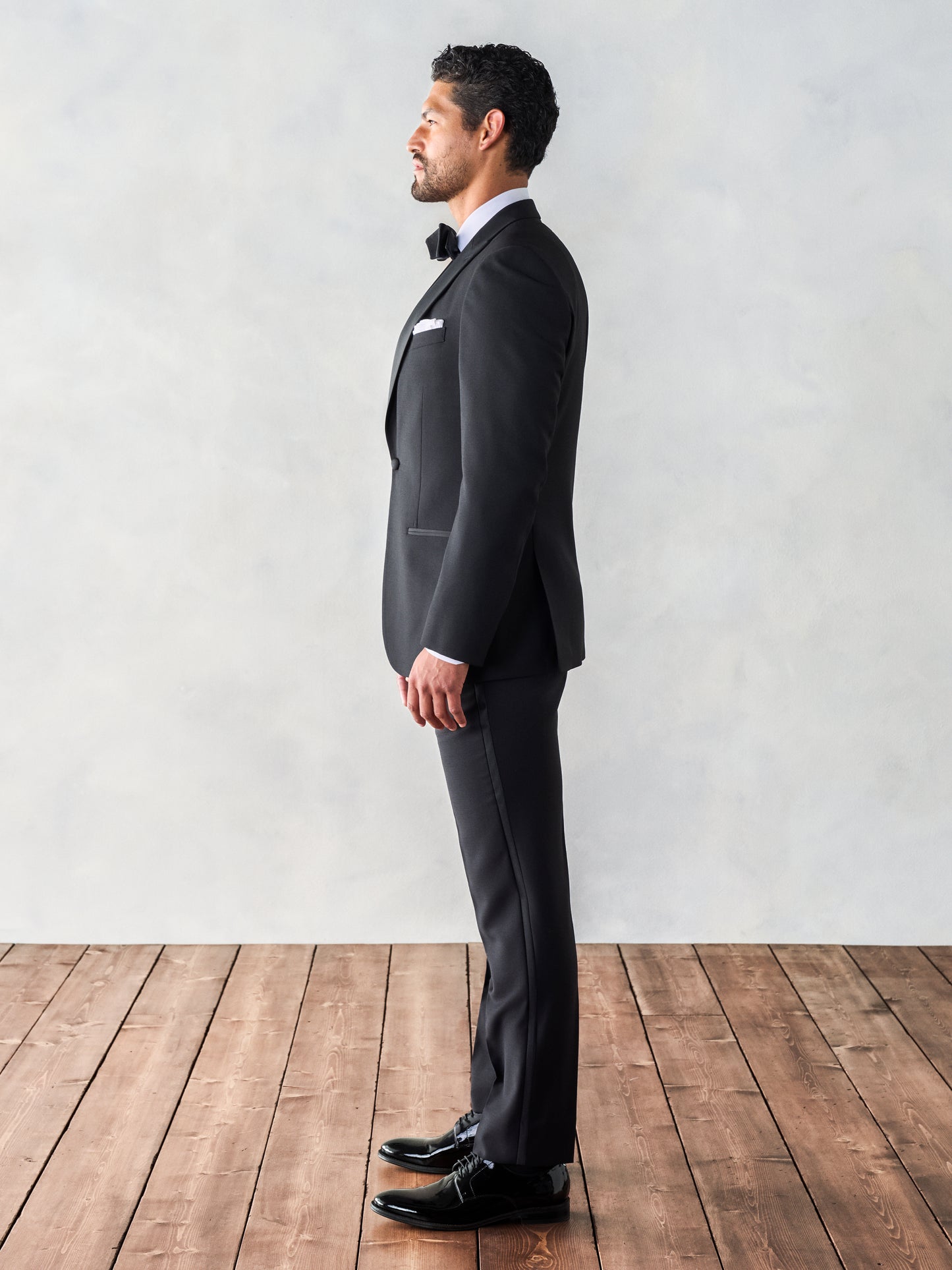 Side view of a man in a black tuxedo, standing straight with his hands by his sides, on wooden floorboards.