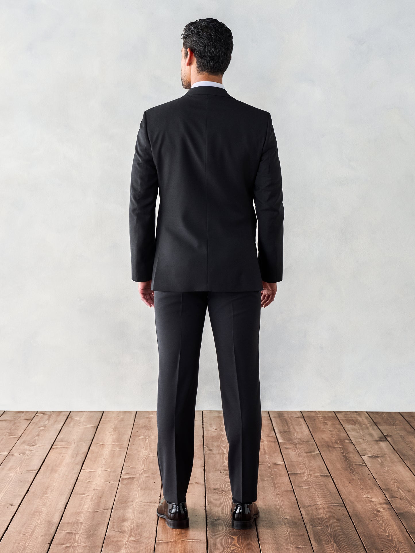 Back view of a man in a black tuxedo, standing on wooden floorboards with a light gray background.