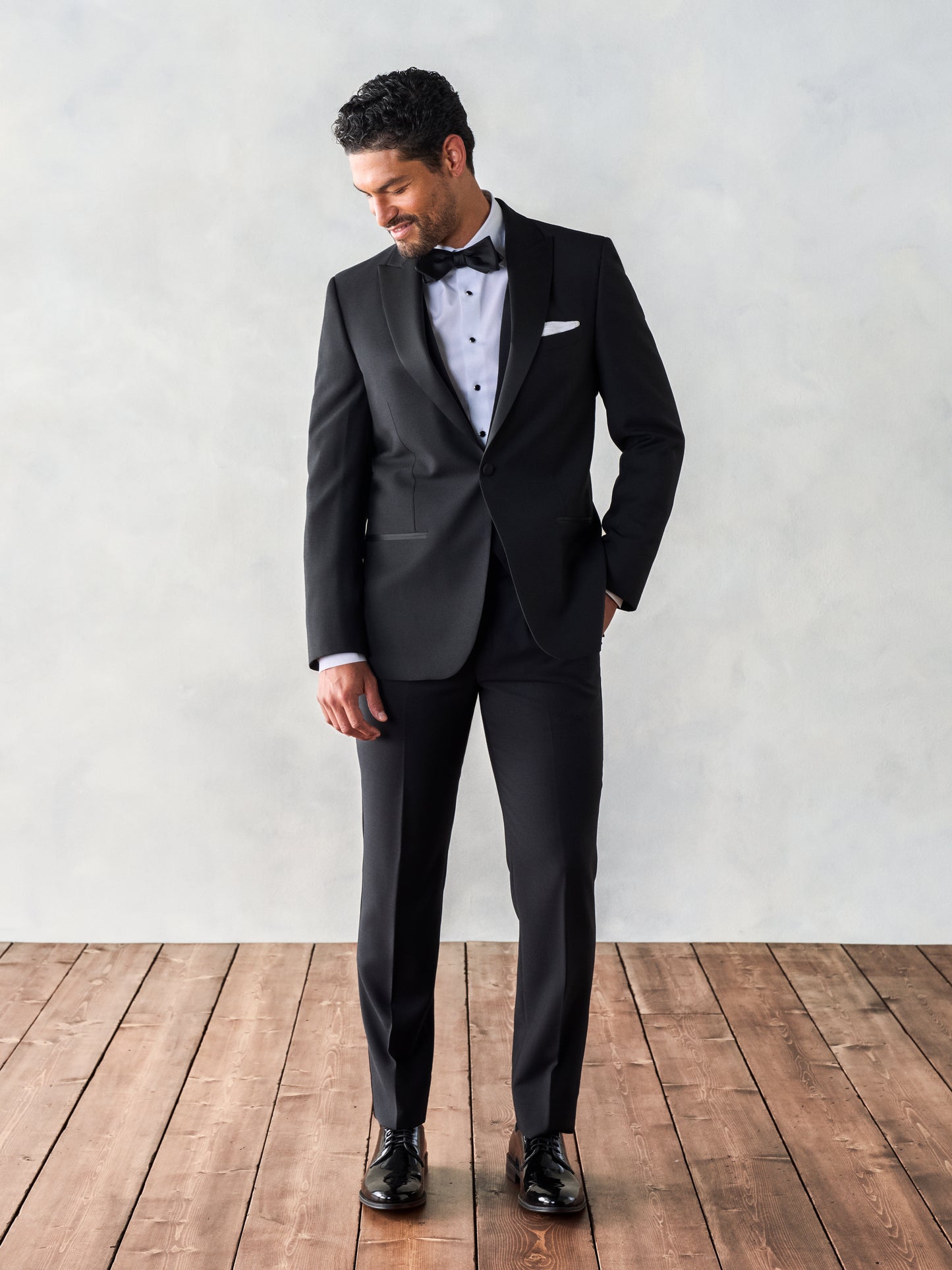 Man wearing a black peaked lapel tuxedo, looking down and smiling, standing on wooden floorboards with a light gray background.