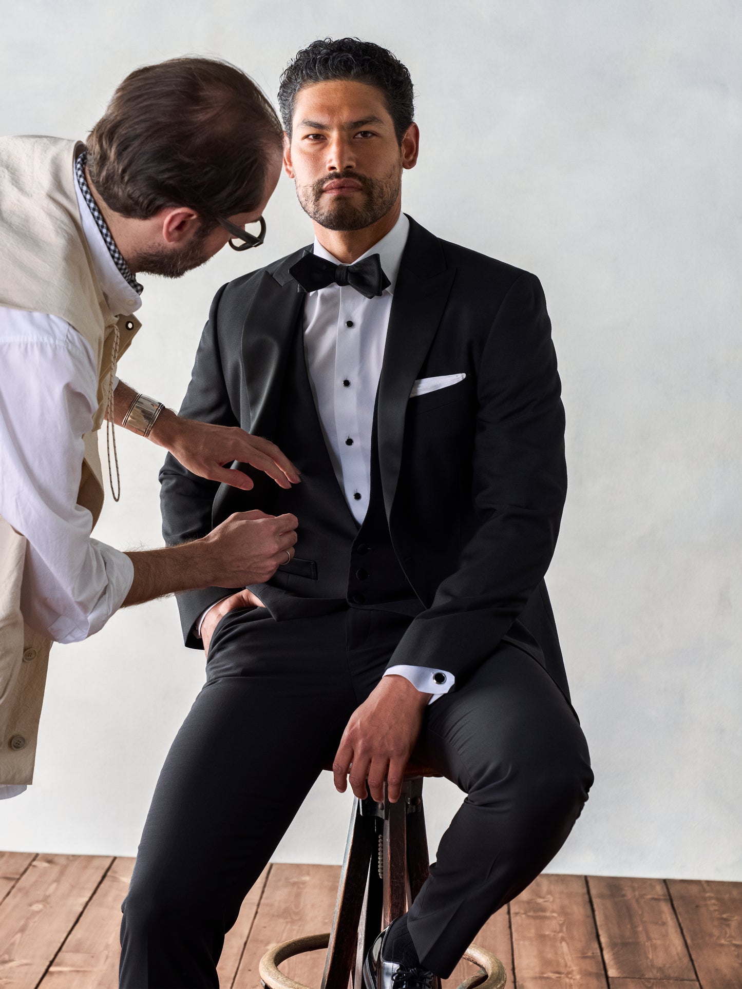 Man sitting on a stool in a black tuxedo with a white shirt and black bow tie, while another man adjusts his jacket.